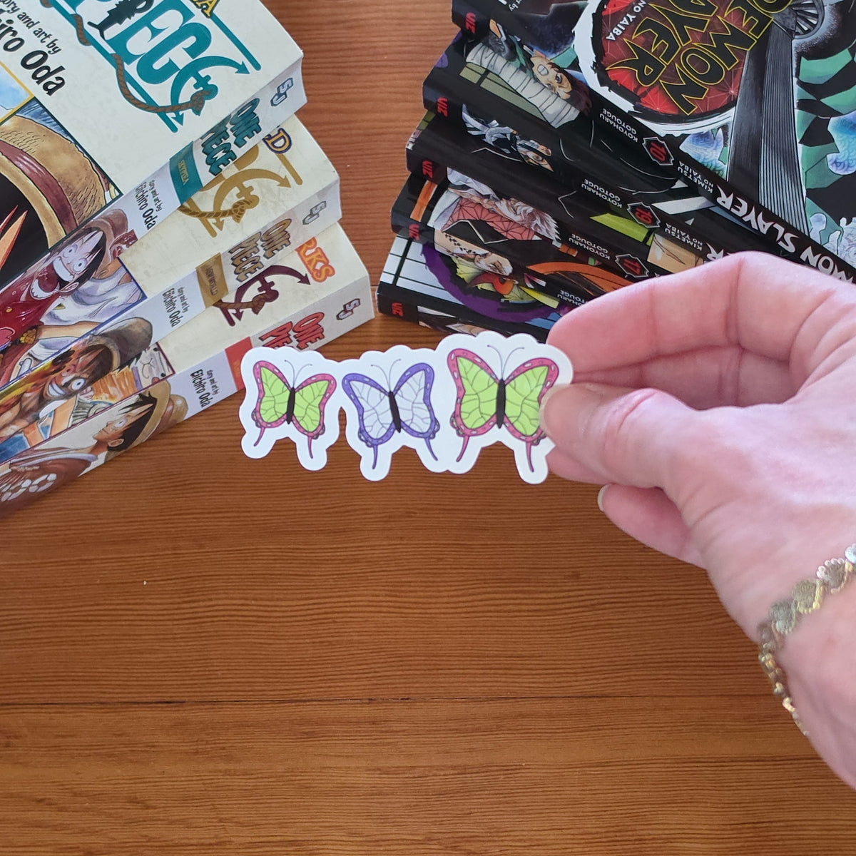 A hand holds a sticker with three small butterflies with white borders and pastel purple, green, and lavender colors above a wooden table, with comic book boxes and a stack of comic books in the background.