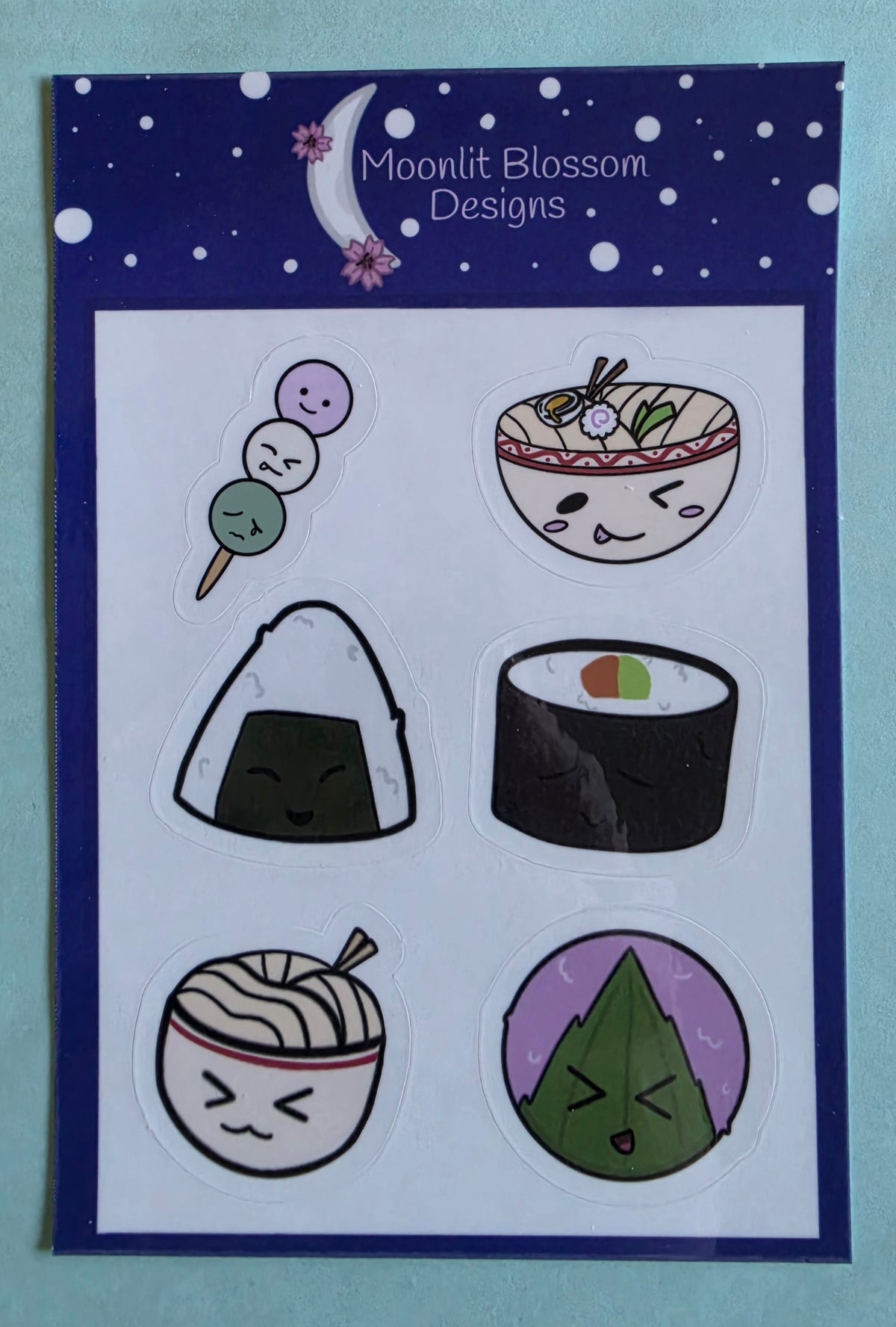A sheet of six cute, cartoon-style anime food stickers with a dark blue border. The card is branded 'Moonlit Blossom Designs' at the top.
