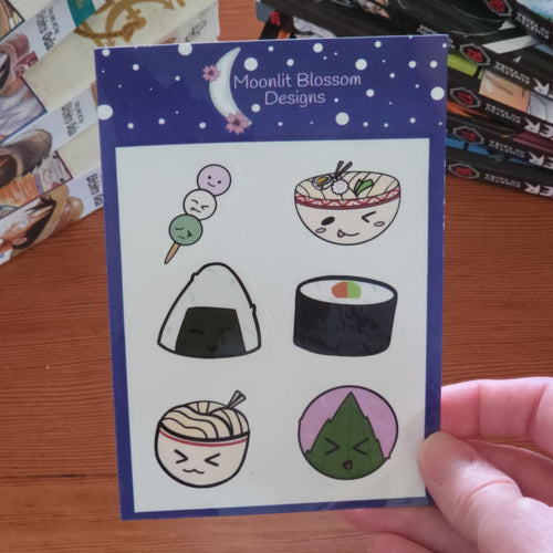 A hand holds a sticker sheet from Moonlit Blossom Designs featuring a sheet of six cute, cartoon-style anime food stickers with a dark blue border. The card is branded 'Moonlit Blossom Designs' at the top. The sheet is held over a wooden desk with books in the background.