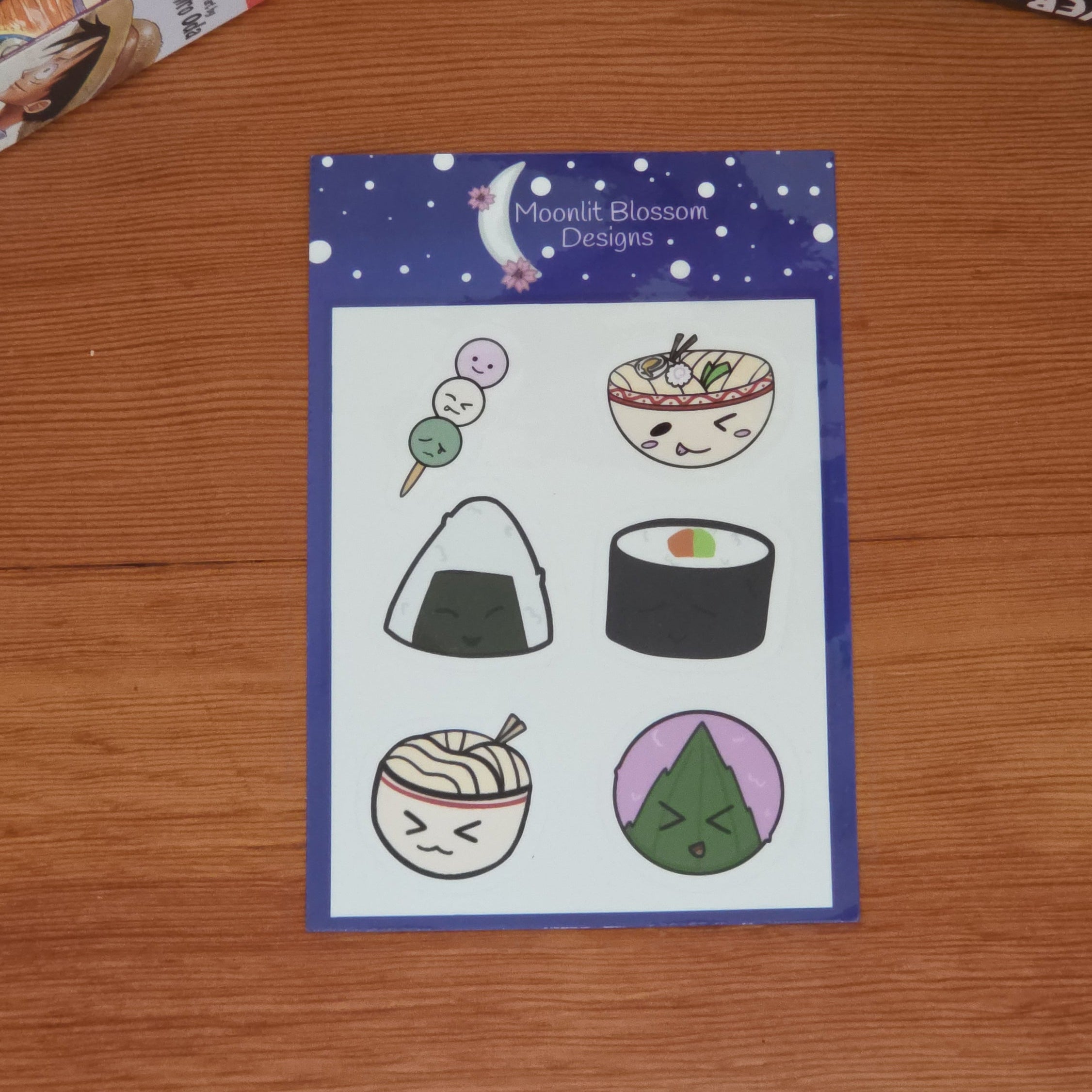 A sheet of cute, hand-drawn Japanese food stickers on a wooden table, surrounded by manga stacks in the background. The sheet features illustrations of sushi, ramen, an onigiri, and a mochi character under a starry night sky header.