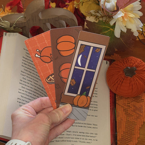 An open book on a wooden table surrounded by autumn-themed decorations with 3 autum-themed bookmarks on the open page.