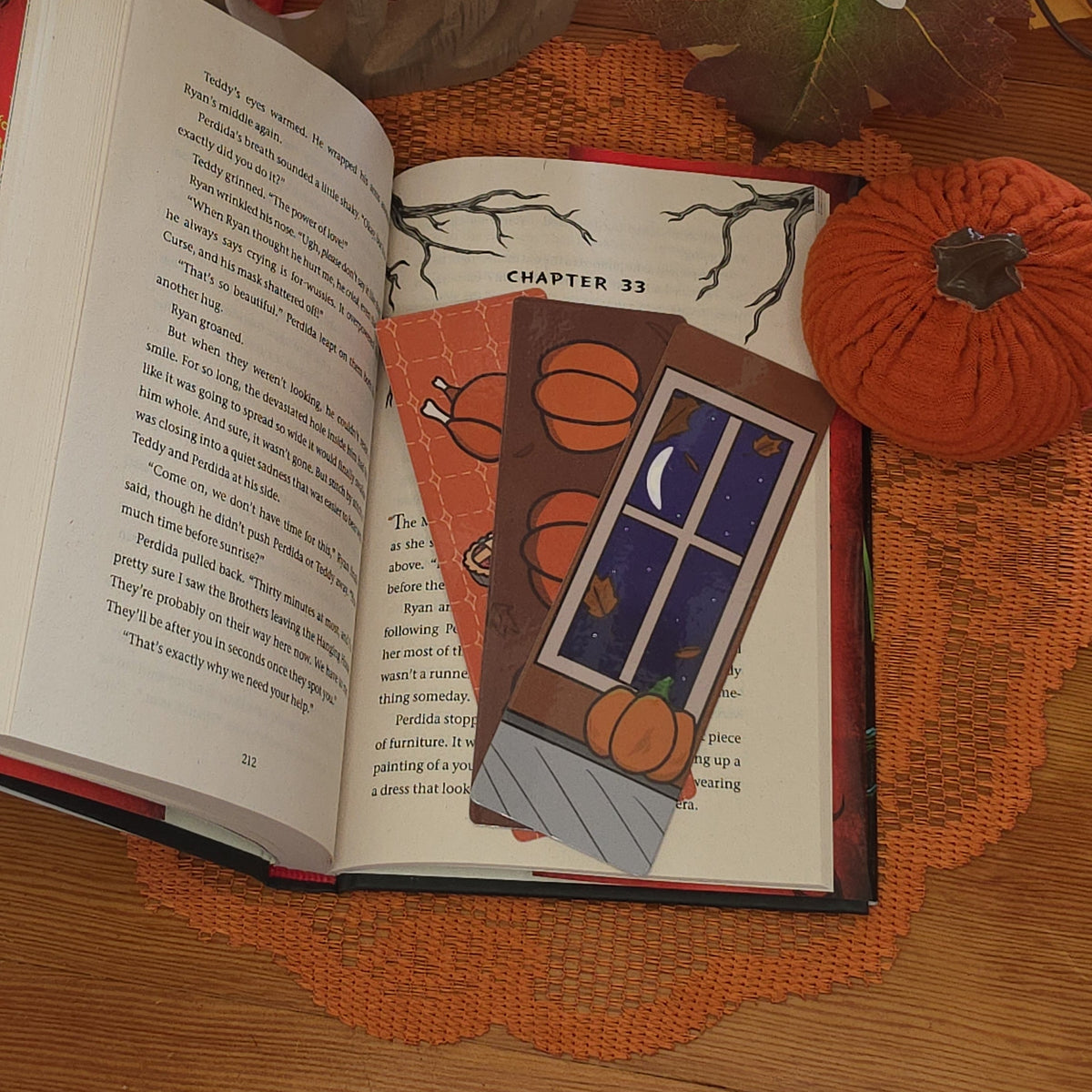 An open book on a wooden table surrounded by autumn-themed decorations with 3 autum-themed bookmarks on the open page.