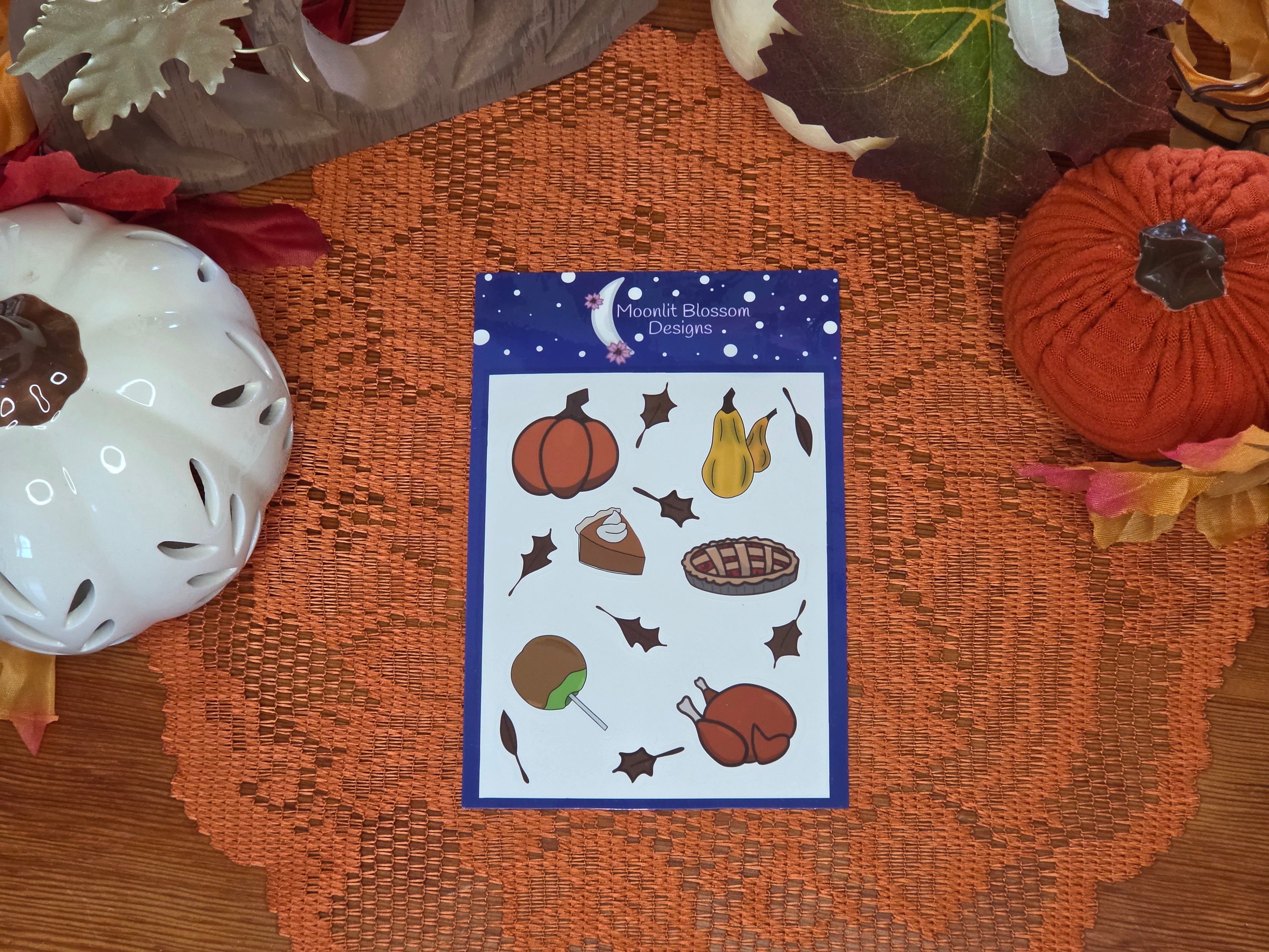 A sheet of autumn-themed stickers from 'Moonlight Blossom Designs' is displayed on an orange lace doily with autum decor in the background. The sheet features illustrations of a pumpkin, gords, a slice of pumpkin pie with whipped cream, a whole cherry pie, a roast turkey, a candied caramel apple, and leaves on a dark blue starry background.