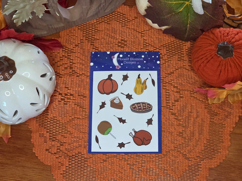 A sheet of autumn-themed stickers from 'Moonlight Blossom Designs' is displayed on an orange lace doily with autum decor in the background. The sheet features illustrations of a pumpkin, gords, a slice of pumpkin pie with whipped cream, a whole cherry pie, a roast turkey, a candied caramel apple, and leaves on a dark blue starry background.