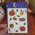 A hand holds a sticker sheet from Moonlit Blossom Designs featuring autumn-themed illustrations like pumpkins, pies, leaves, androast turkey on a white background with a blue border, set against an orange lace doily and fall decorations.