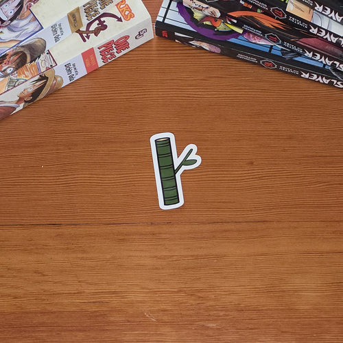 A small die-cut sticker shaped like a green bamboo shoot with a white border lies on a wooden tabletop. Manga books are stacked on both sides of the table.