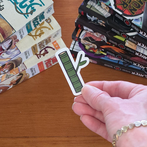 A hand holding a small die-cut sticker shaped like a green bamboo stalk illustration with black outlines and white borders, photographed over a wooden tabletop with manga books in the background.