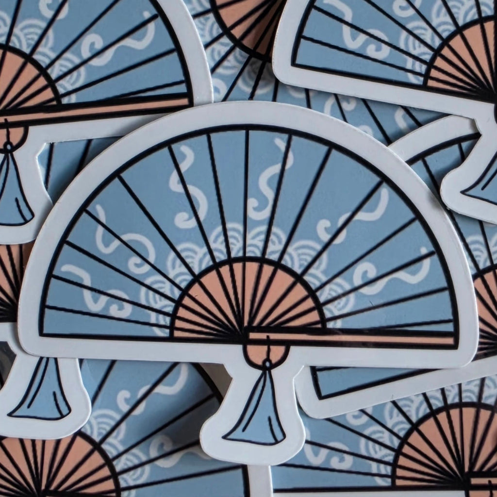 A close-up of multiple die-cut stickers featuring a stylized folding fan design. The fan has a light blue background with black line details, peach-pink curved segments, and white outlines, layered over a matching patterned surface.