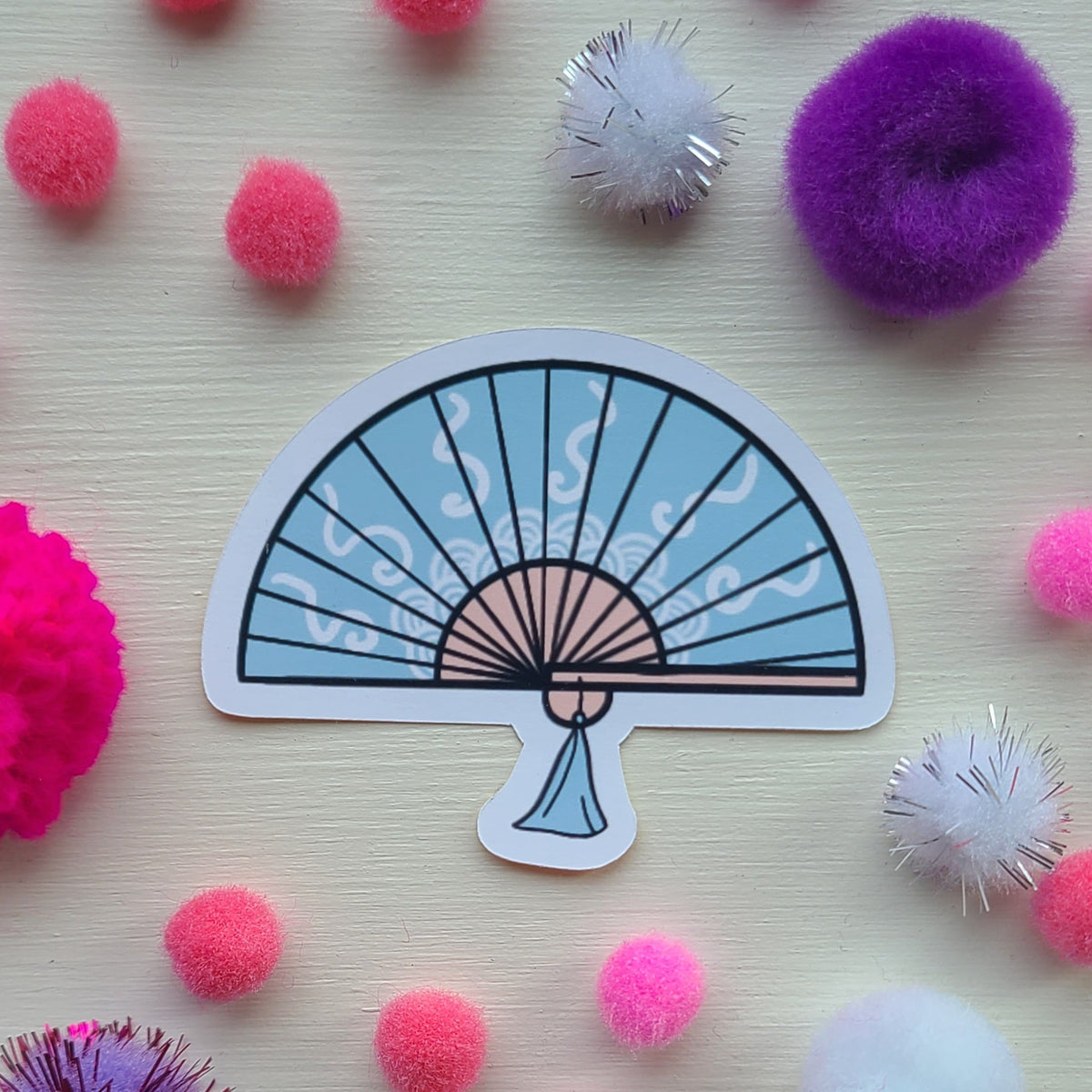 Sticker of a blue fan design on a light wooden surface with colorful pom-poms.
