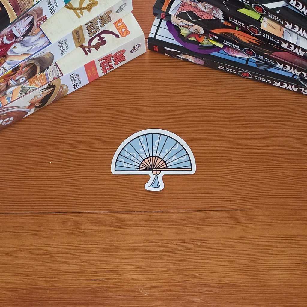 A small die-cut sticker shaped like a folding fan with a blue and white striped pattern and a beige center, placed on a wooden tabletop. Manga books are stacked in the top-left and top-right corners.