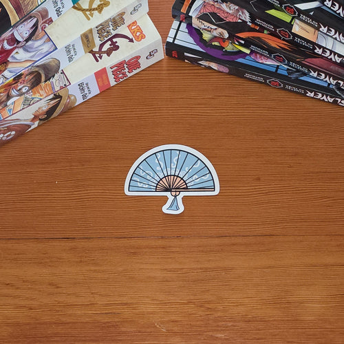 A small die-cut sticker shaped like a folding fan with a blue and white striped pattern and a beige center, placed on a wooden tabletop. Manga books are stacked in the top-left and top-right corners.