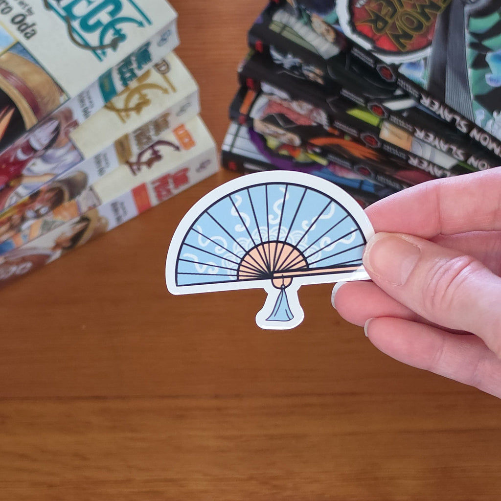A hand holding a die-cut sticker shaped like a folding fan with a light blue and white striped pattern and a small white accent, set against a wooden table with manga books in the background.