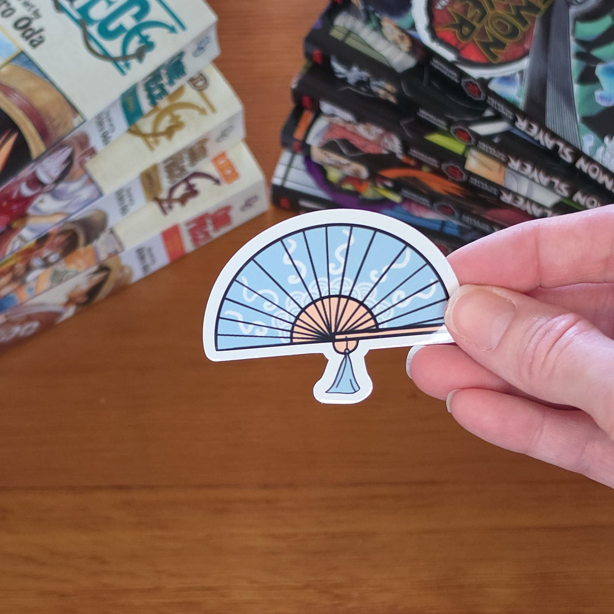A hand holding a die-cut sticker shaped like a folding fan with a light blue and white striped pattern and a small white accent, set against a wooden table with manga books in the background.