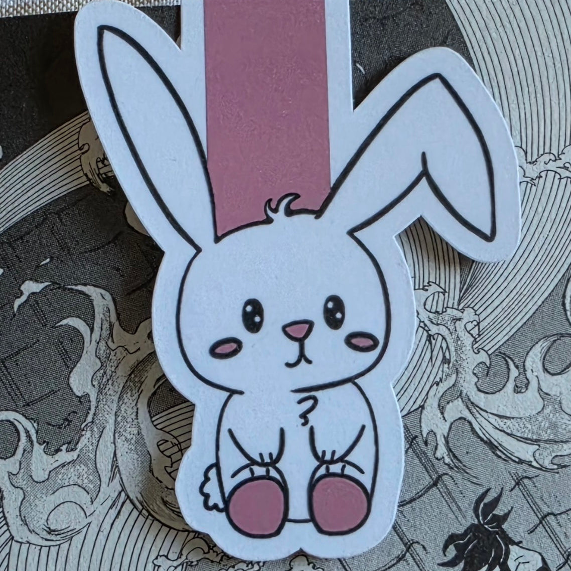 A cartoon bunny magnetic bookmark with a white body, black outlines, and dusty pink accents, shown against an open page of a manga book background.