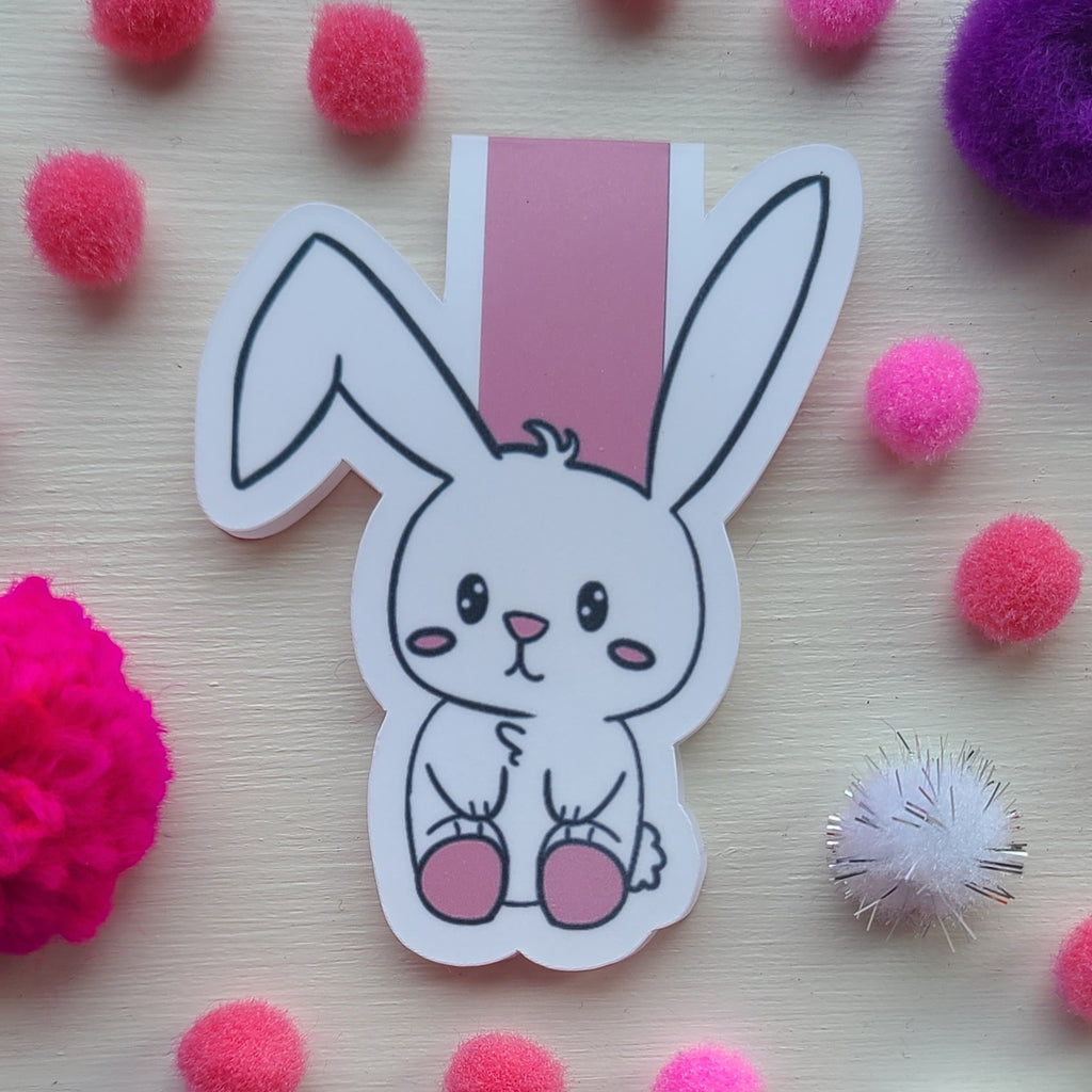 Cartoon bunny magnetic bookmark on a light wooden background with colorful pom-poms.