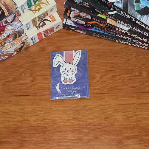 A small blue card featuring a white cartoon bunny magnetic bookmark is placed on a wooden table, with manga books stacked on either side.