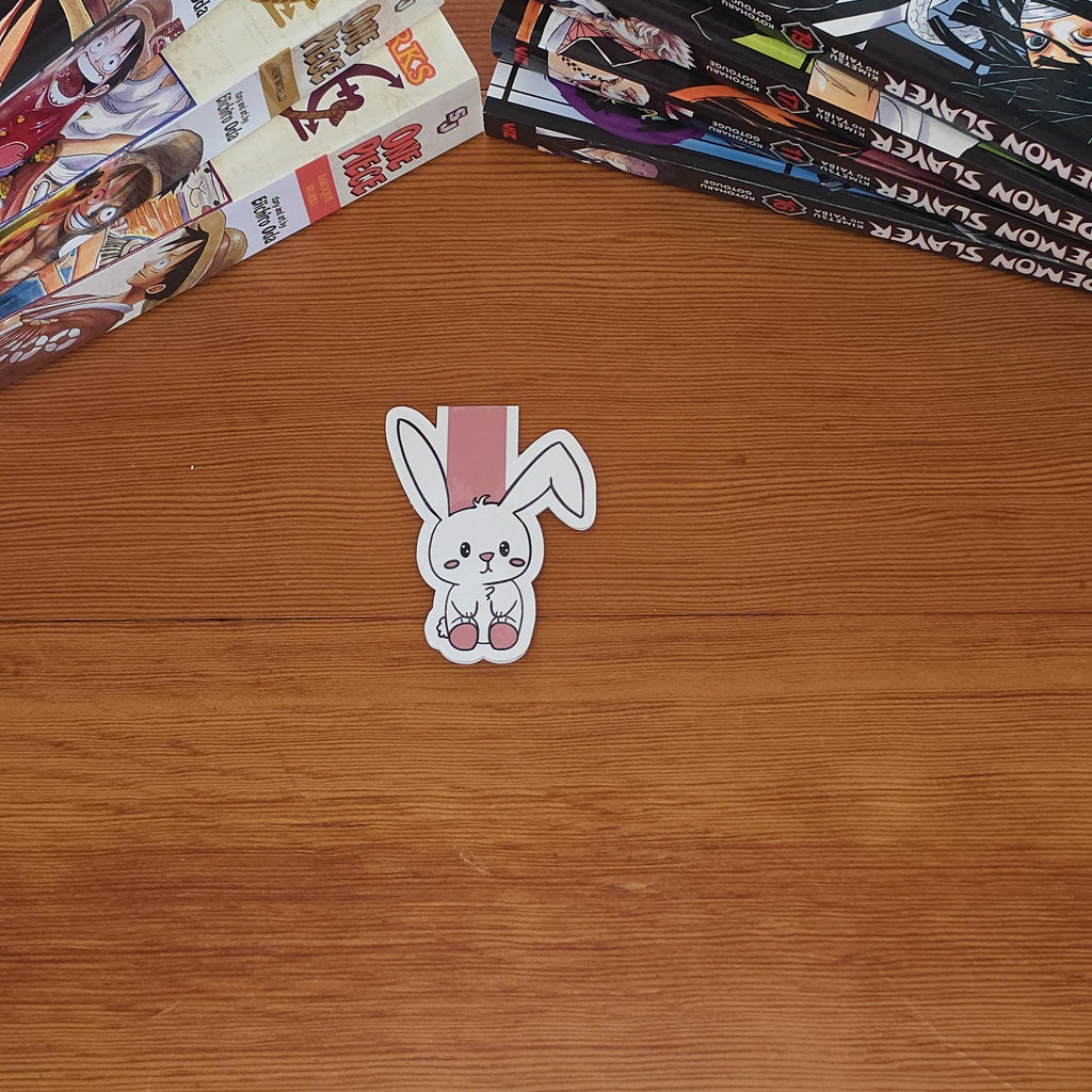 A small white bunny magnetic bookmark with pink and black details sits on a wooden tabletop, with stacks of manga books visible in the background.