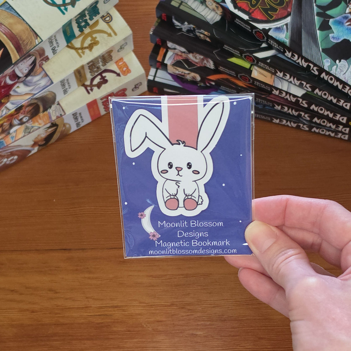 A hand holding a square magnetic bookmark featuring a cartoon bunny on a starry sky background, with manga books stacked on a wooden table in the background.