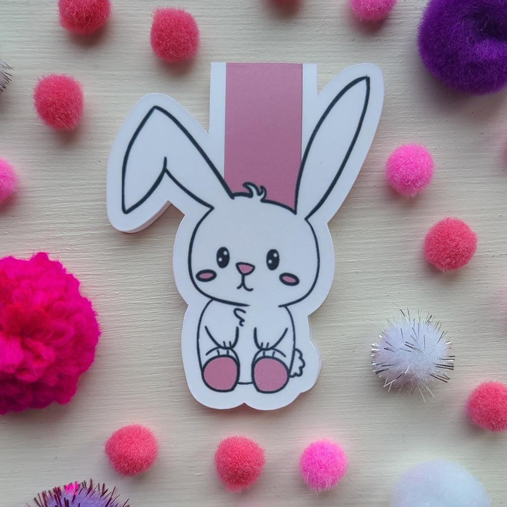 A cute cartoon bunny sticker with pink feet, placed on a light wood surface surrounded by colorful pom-poms.