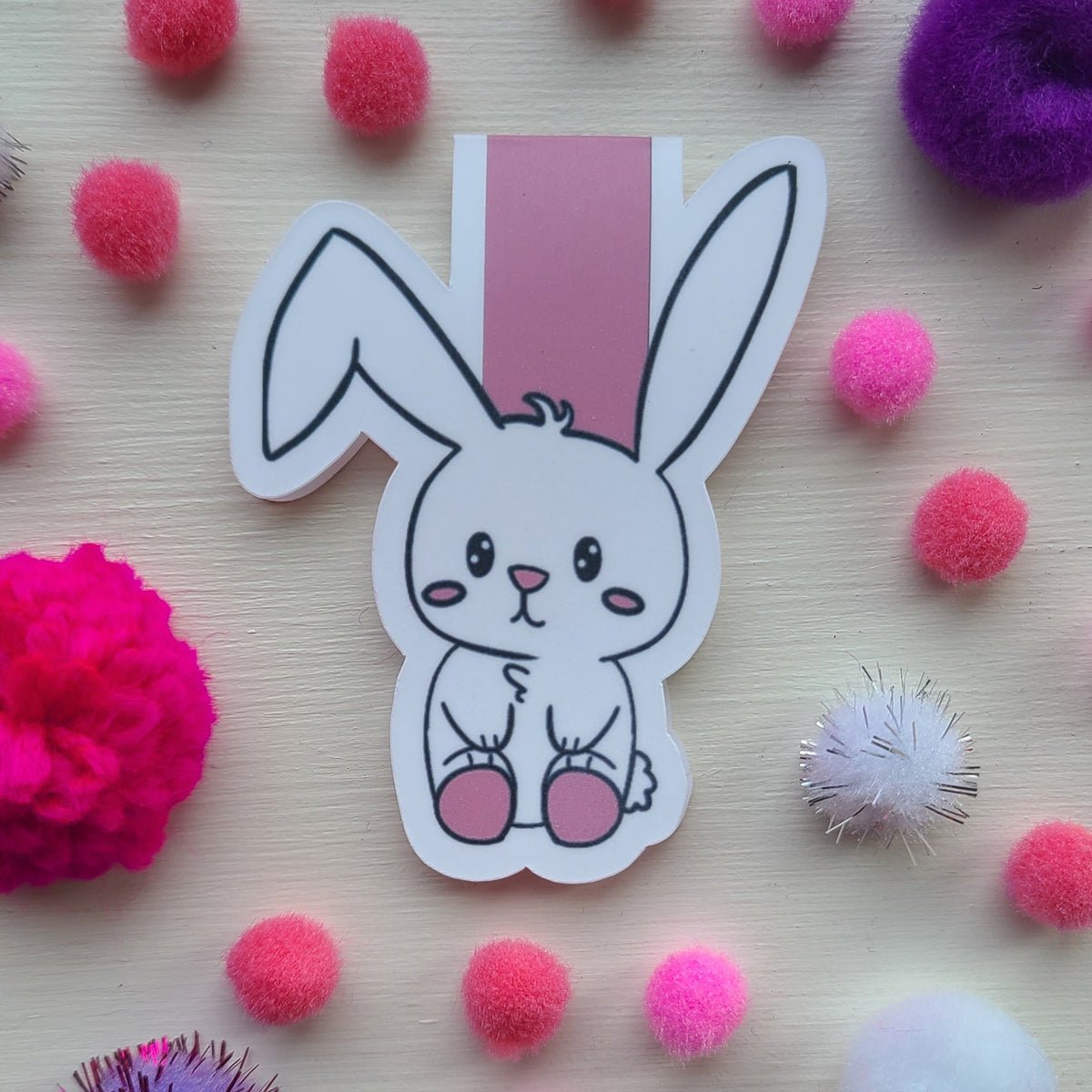 A cute cartoon bunny sticker with pink feet, placed on a light wood surface surrounded by colorful pom-poms.