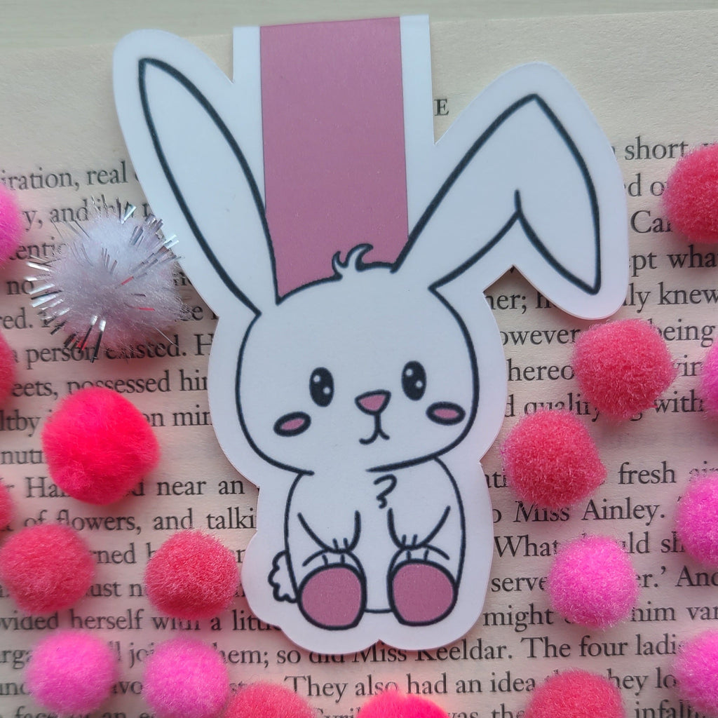A white bunny-shaped bookmark with long black-outlined ears, a pink nose, and pink feet, placed on top of an open book and surrounded by bright pink pom-poms.