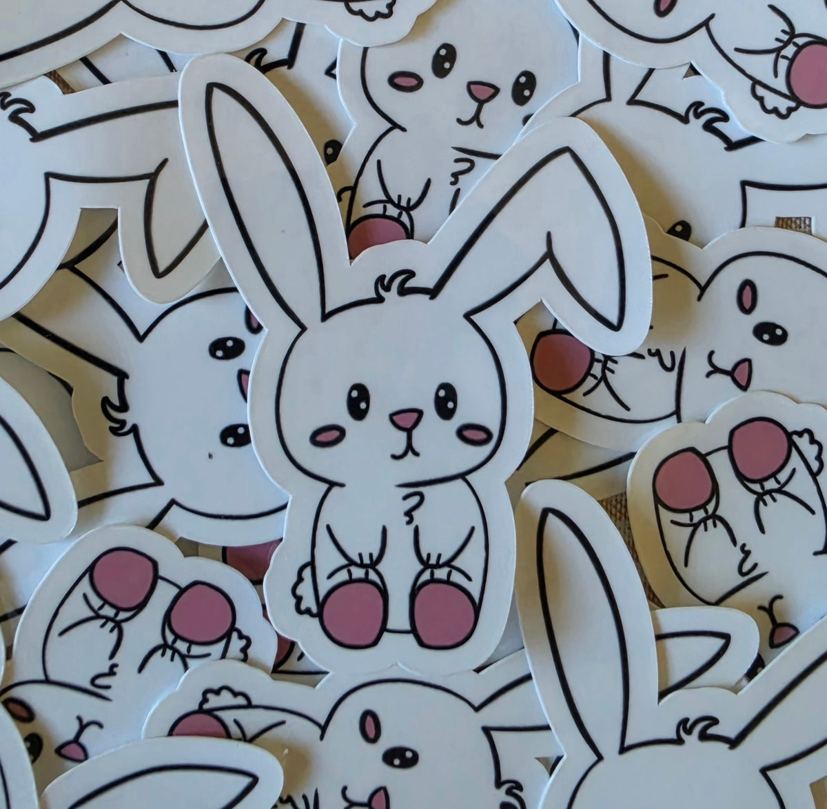 A pile of white bunny-shaped stickers with black outlines and pink accents, featuring cute cartoon bunnies with pink cheeks, pink noses, and pink feet.