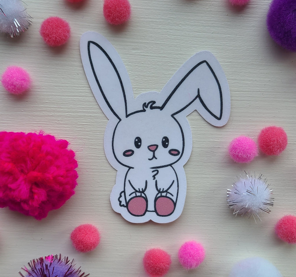 Cartoon bunny sticker surrounded by colorful pom-poms on a light wooden surface