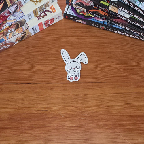 A small white bunny sticker with black outlines and pink details sits on a wooden tabletop, with stacks of manga books in the background.