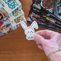 A hand holds a small white bunny sticker with pink feet over a wooden table, with stacks of manga books in the background.