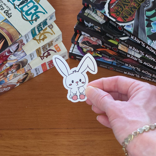 A hand holds a small white bunny sticker with pink feet over a wooden table, with stacks of manga books in the background.