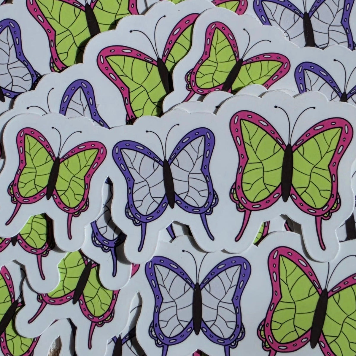 A pile of die-cut stickers featuring colorful butterfly designs on a white background. The butterflies are shown in multiple colorways, including lime green with pink outlines, purple with gray outlines, and green with purple outlines.