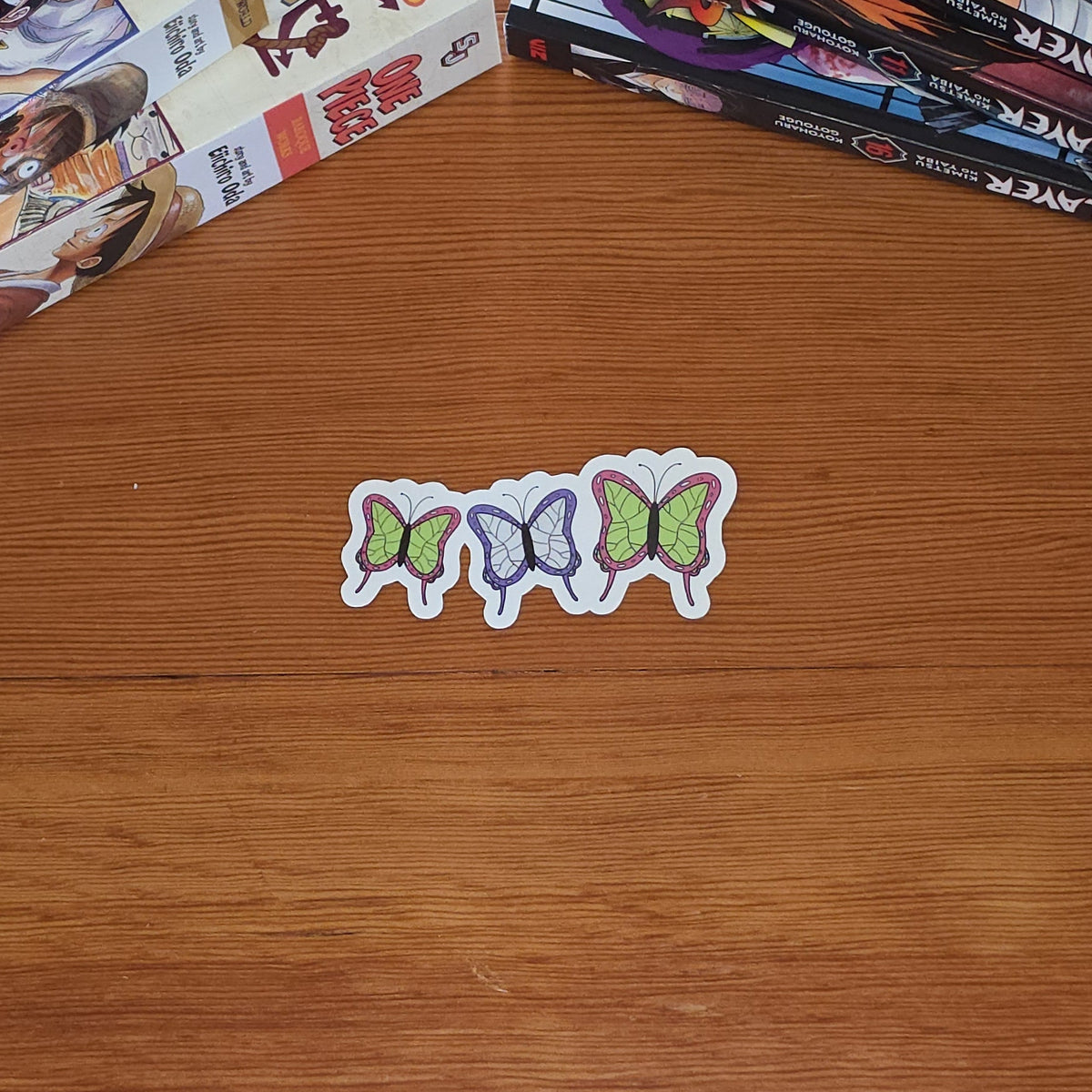 One sticker with three small butterflies in pink, purple, and green are arranged on a wooden tabletop, with stacks of manga books in the background.
