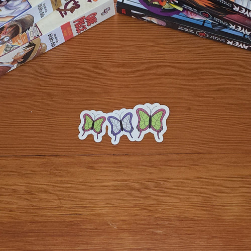 One sticker with three small butterflies in pink, purple, and green are arranged on a wooden tabletop, with stacks of manga books in the background.