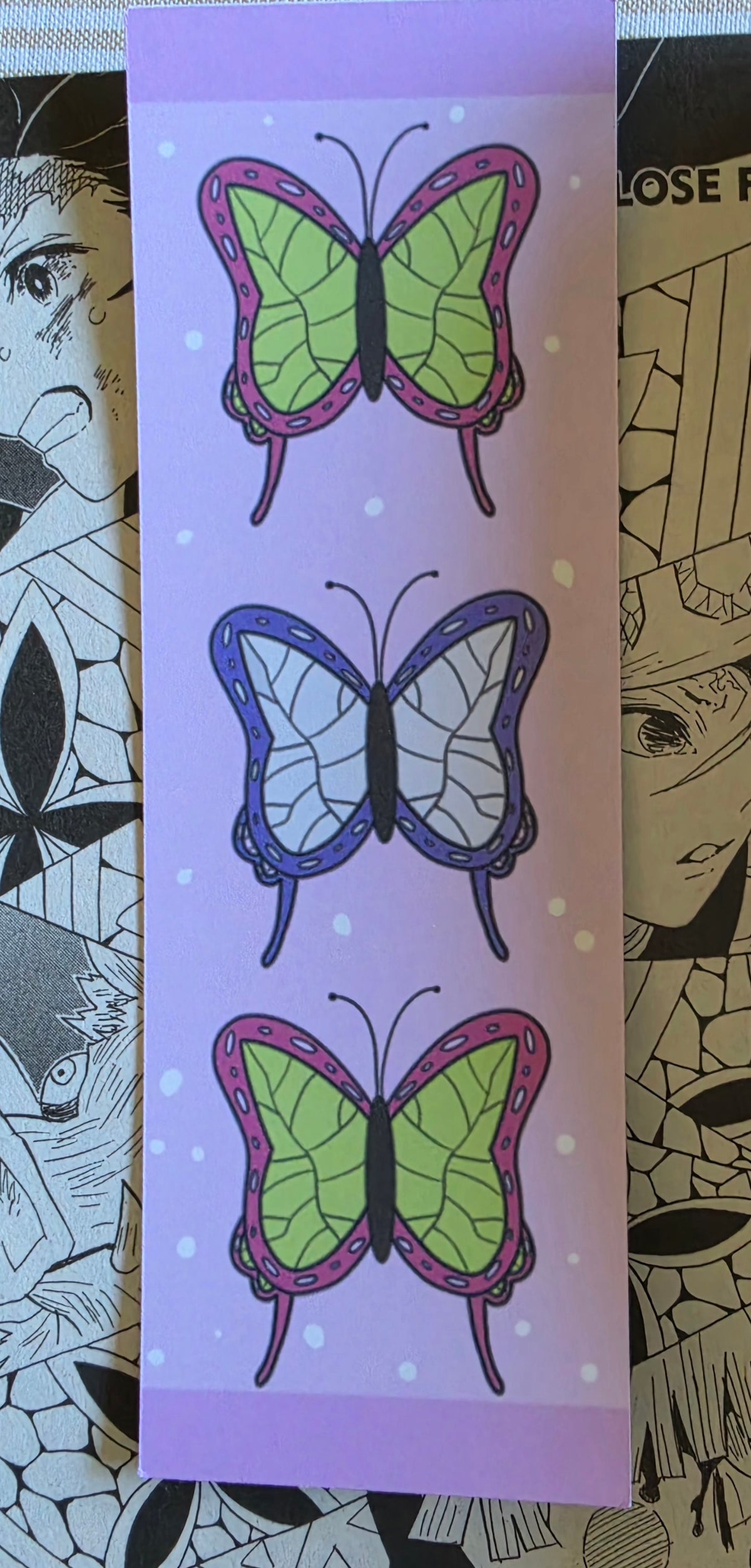 A light pink bookmark featuring three vertical rows of illustrated butterflies in green, purple, and pink, set against a background of anime-style line art.