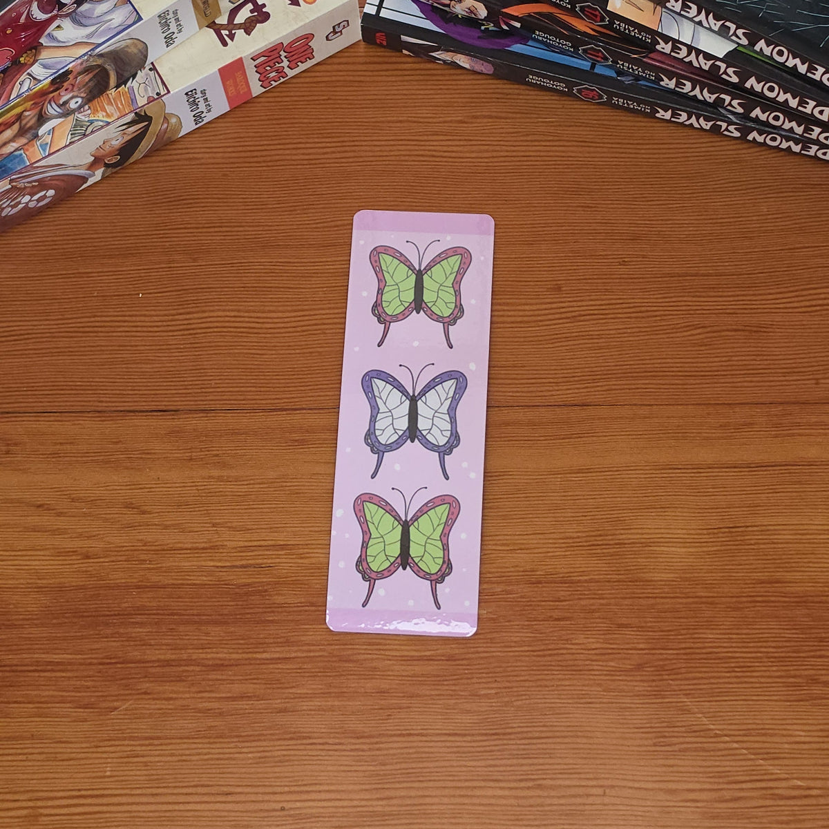 A light pink bookmark featuring three vertical rows of illustrated butterflies in green, purple, and pink, placed on a wooden tabletop with manga books in the background.
