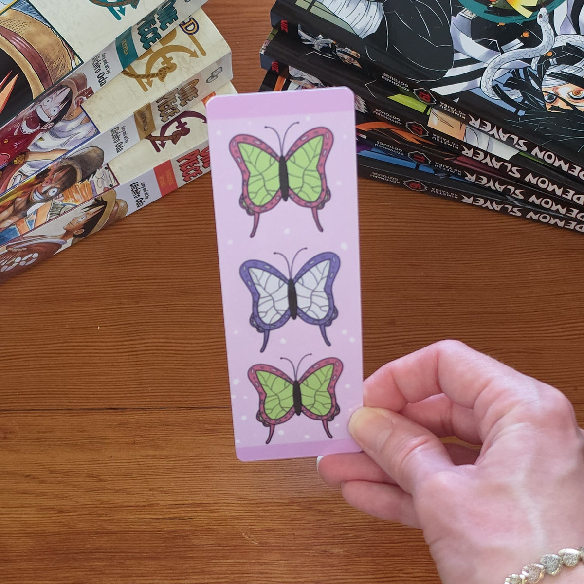 A hand holds a light pink bookmark featuring three vertical rows of illustrated butterflies in green, purple, and pink against a wooden table background with manga books.