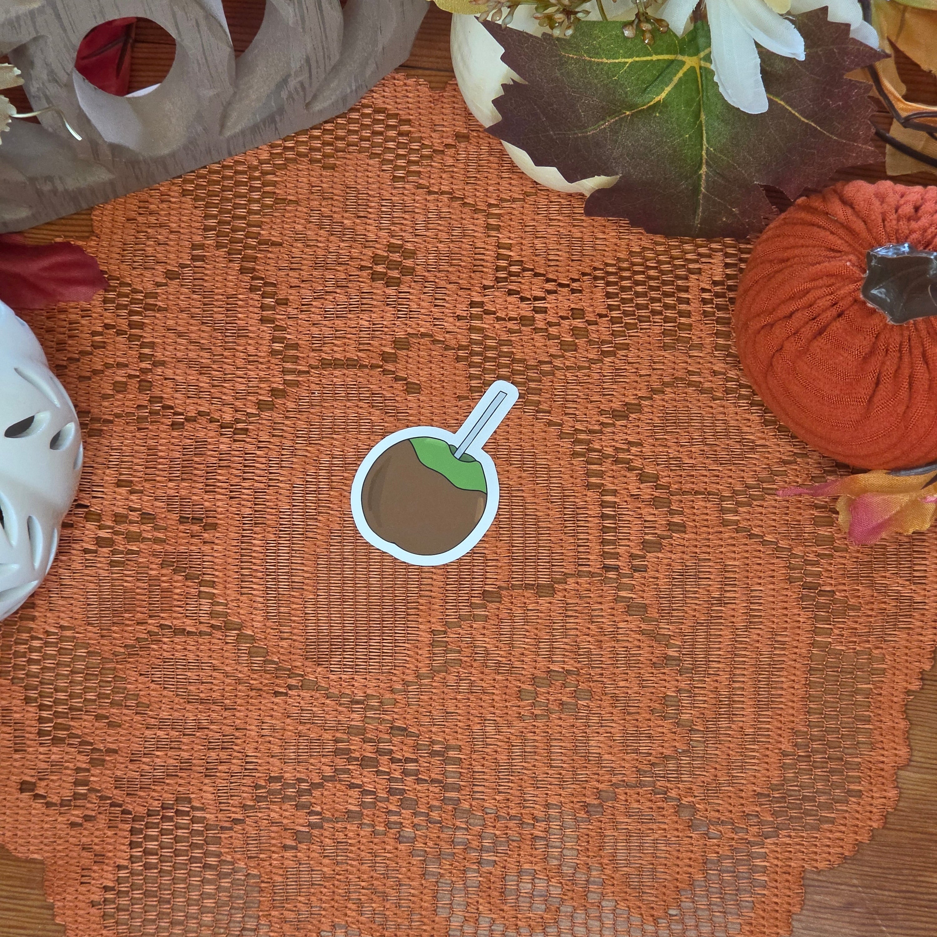 A sticker in the shape of a caramel apple sitting on an orange doily with autumn decor in the background.
