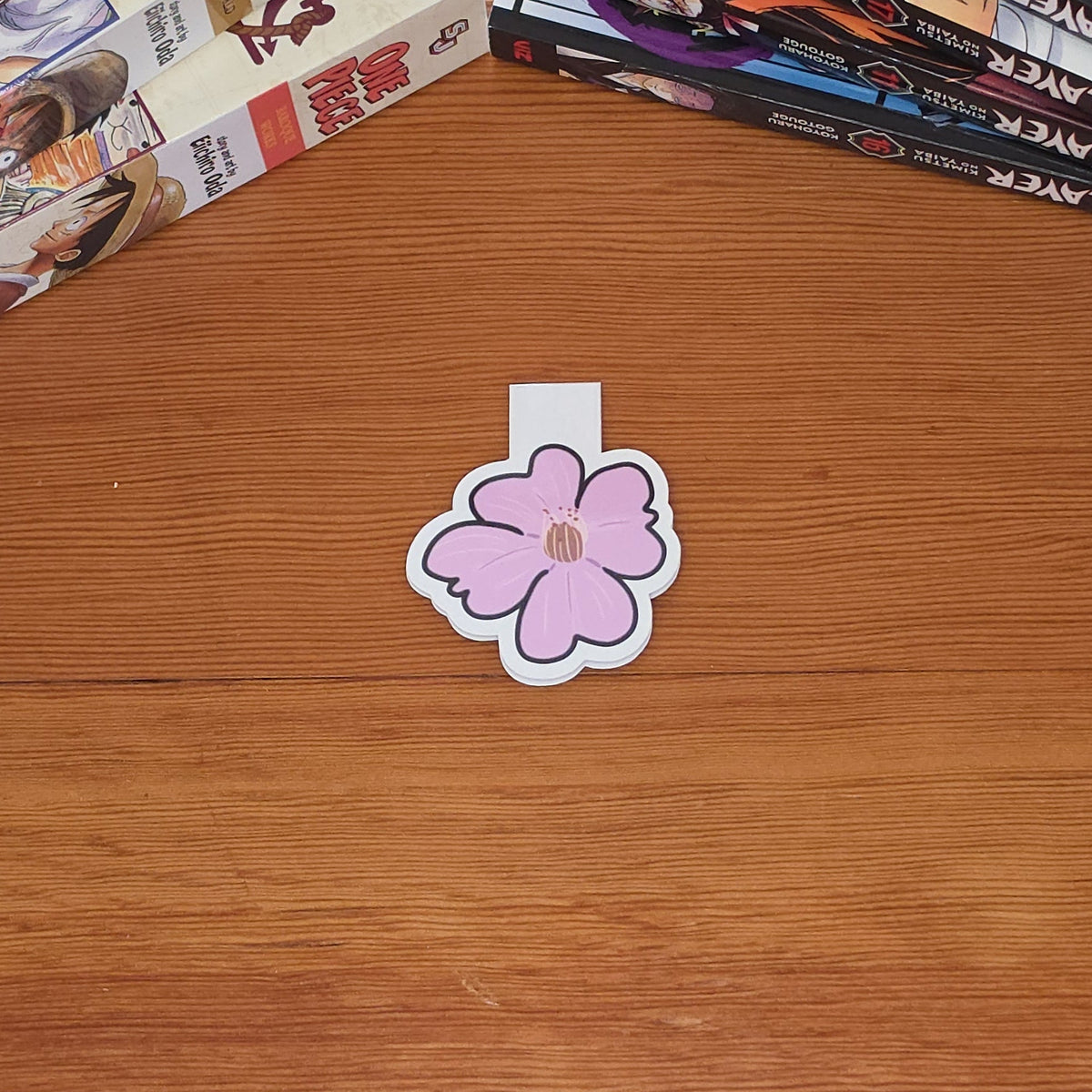 A magnetic bookmark featuring a stylized pink cherry blossom (sakura) flower with a brown center and black outline sits on a wooden tabletop. Manga books are stacked in the background on both sides.