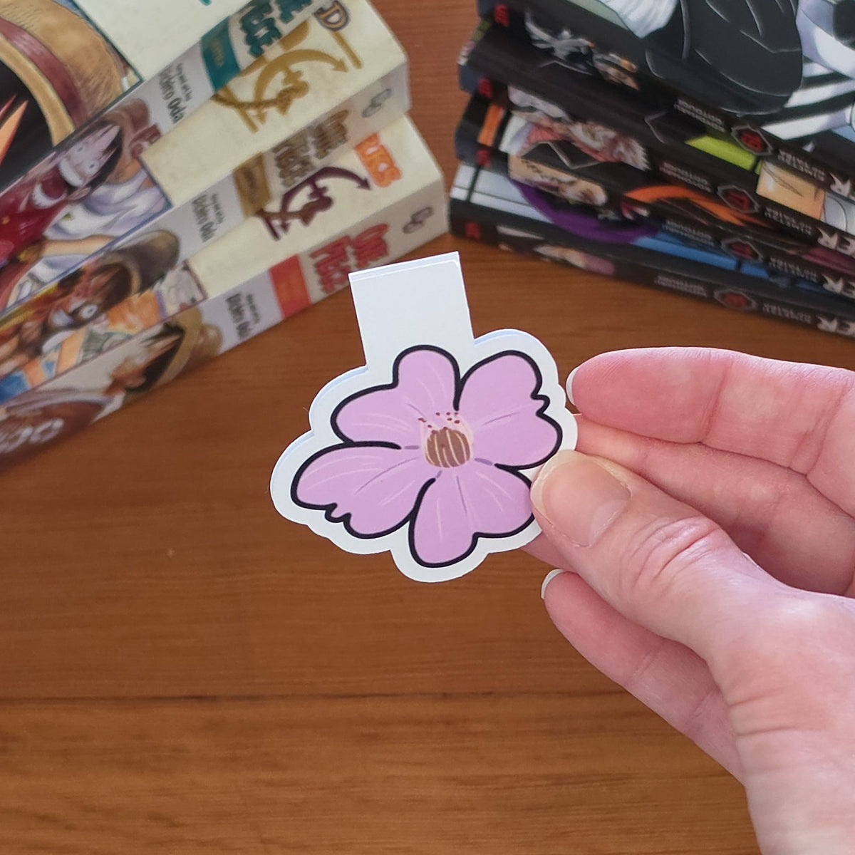 A hand holding a magnetic bookmark featuring a stylized pink cherry blossom (sakura) flower with a brown center and black outline over a wooden table, with manga books visible in the background.