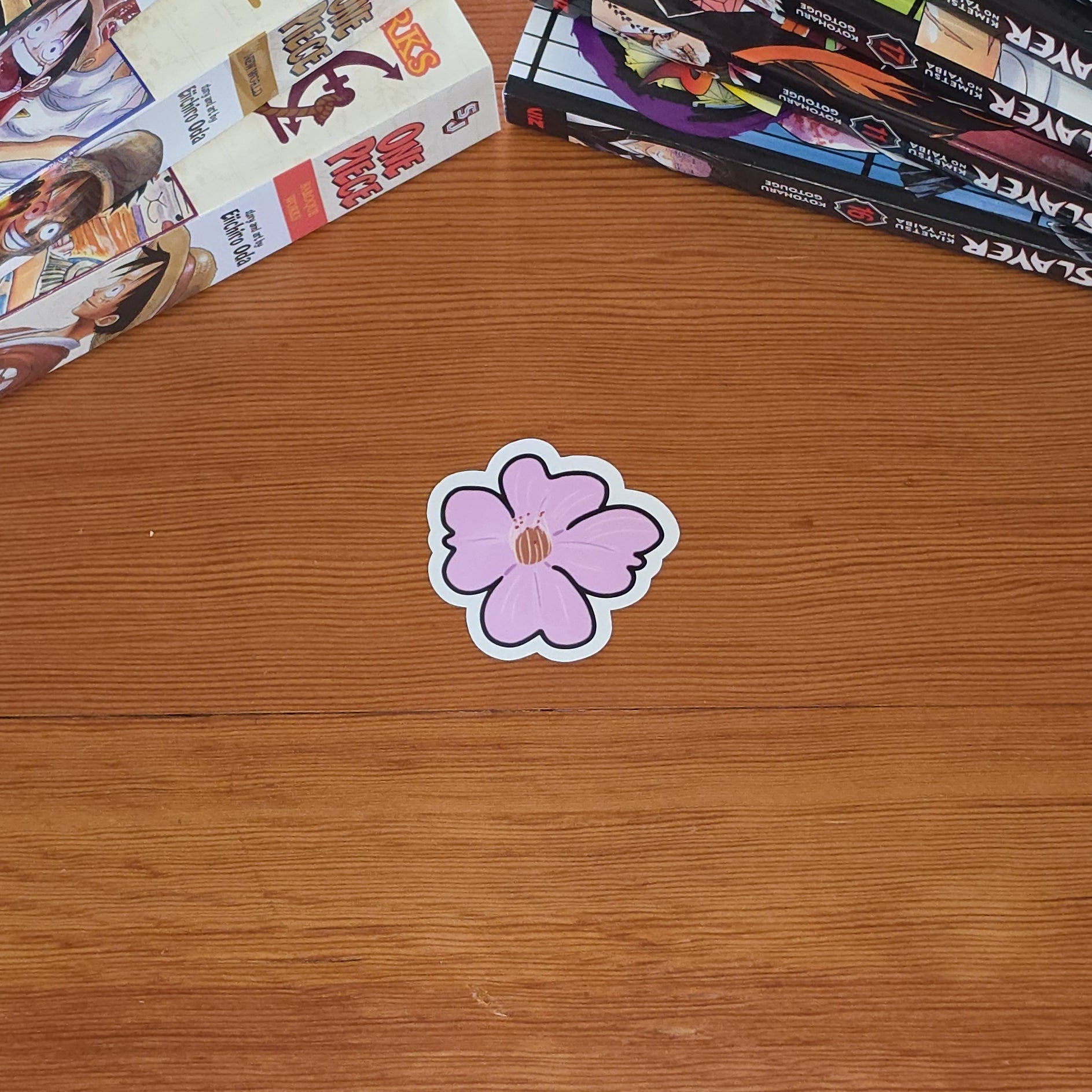 A small pink cherry blossom (sakura) flower shaped sticker with a white border lies on a wooden tabletop, with manga books stacked on both sides of the frame.