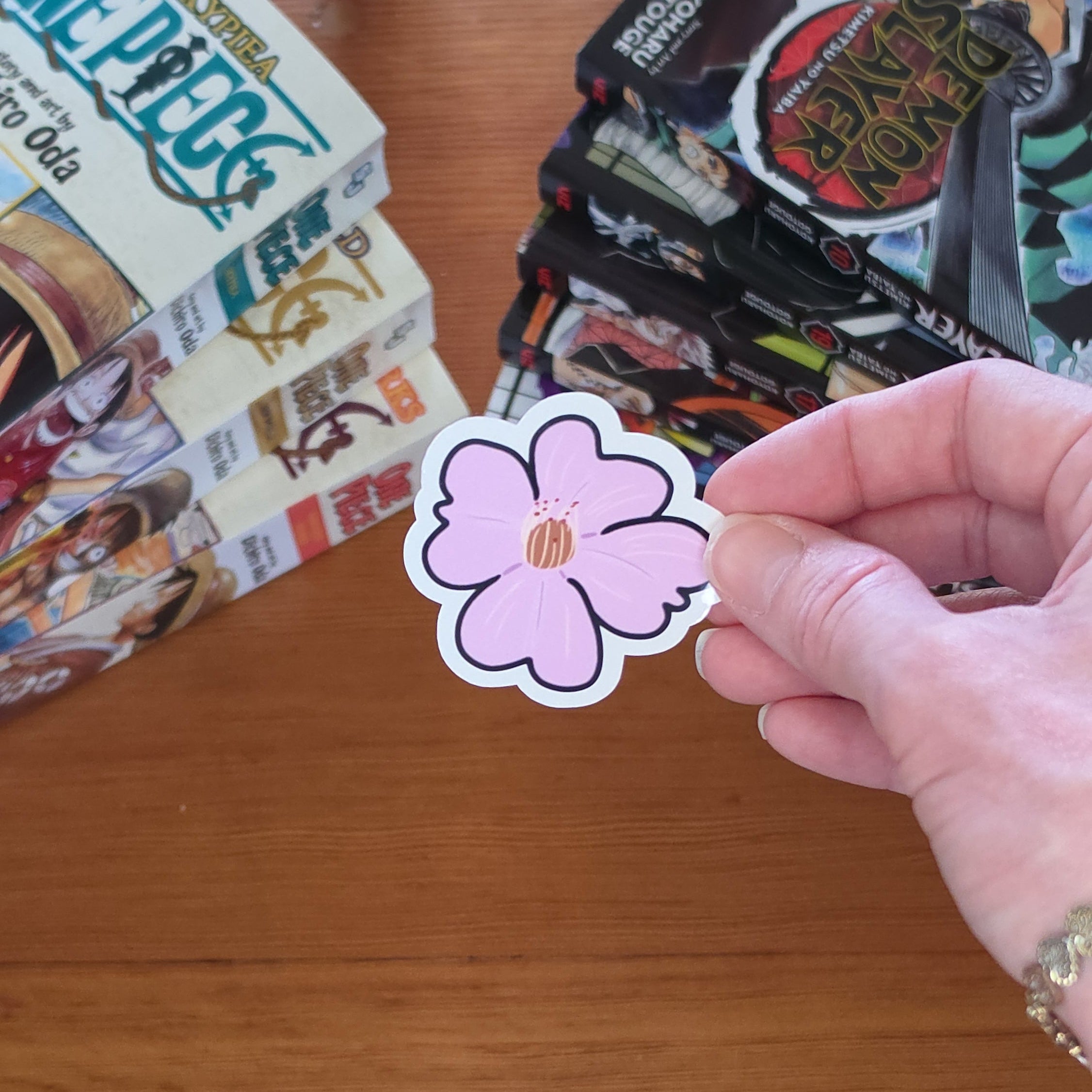 A hand holding a pink cherry blossom sakura flower shaped sticker with a white border and a small brown center, set against a wooden tabletop with manga books in the background.