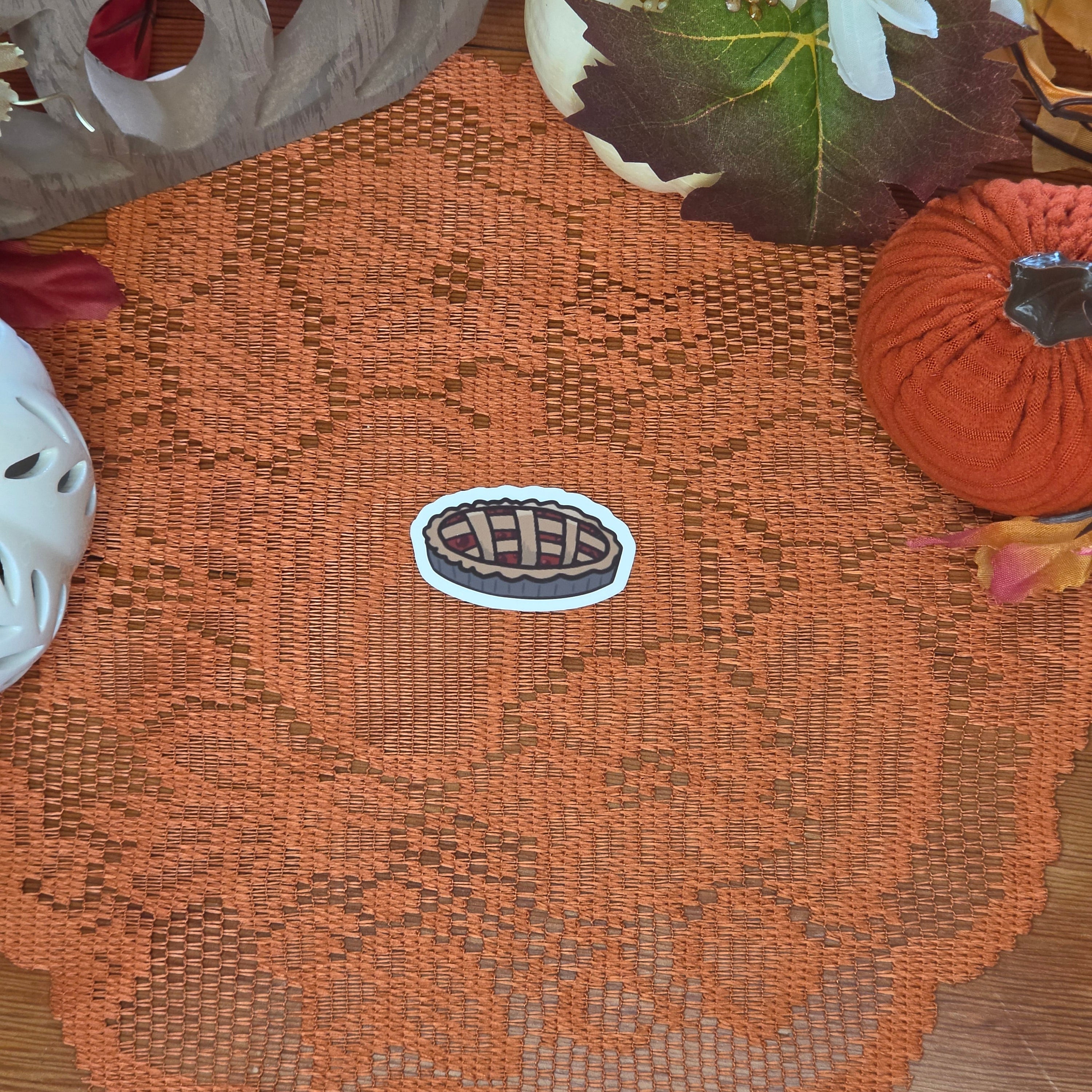 A sticker in the shape of a cherry pie sitting on an orange doily with autumn decor in the background.