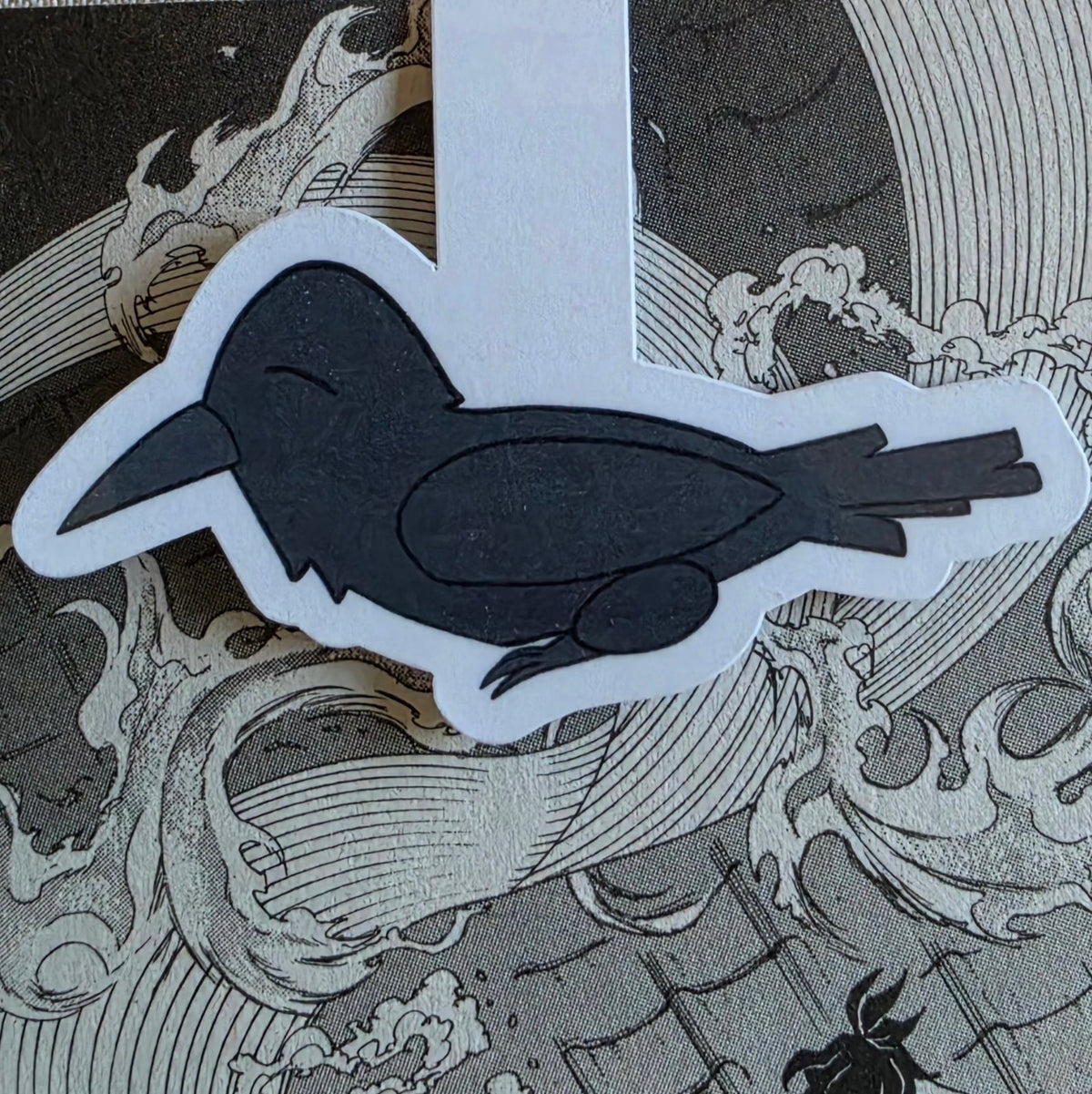 A die-cut sticker of a black raven (crow) resting on a gray and white manga illustration background featuring waves.