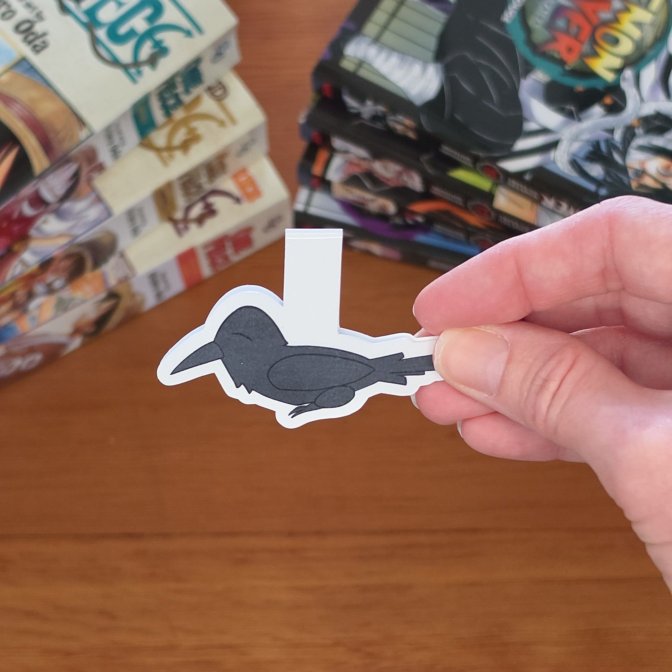 A hand holding a magnetic bookmark of a black raven (crow), set against a wooden tabletop with blurred manga books in the background.