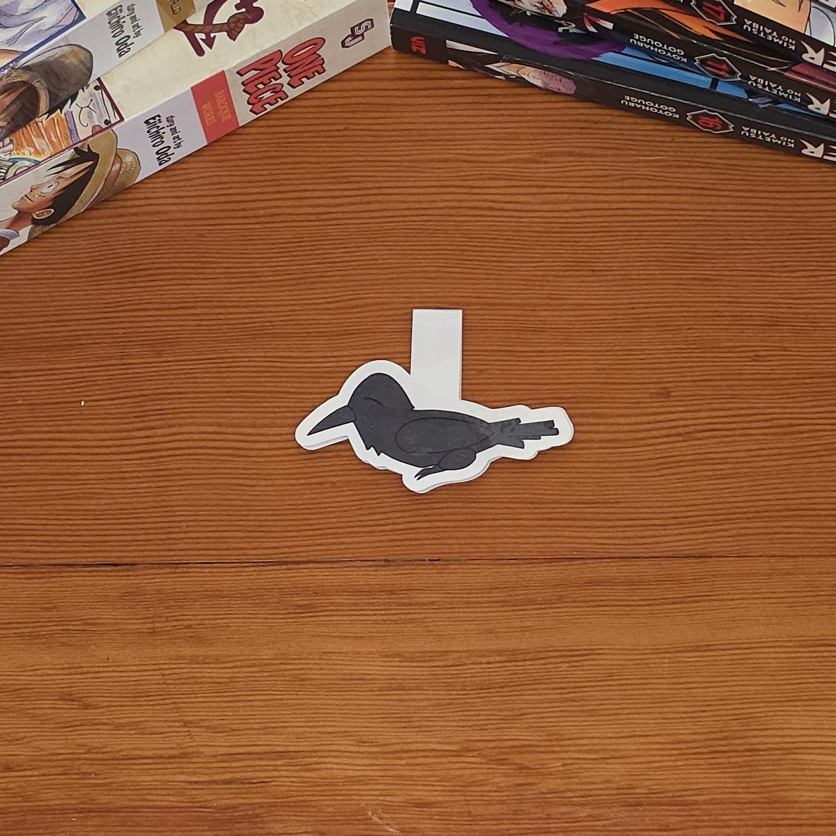 A small magnetic bookmark in the shape of a crow is placed on a wooden tabletop, with stacks of manga books on both sides.
