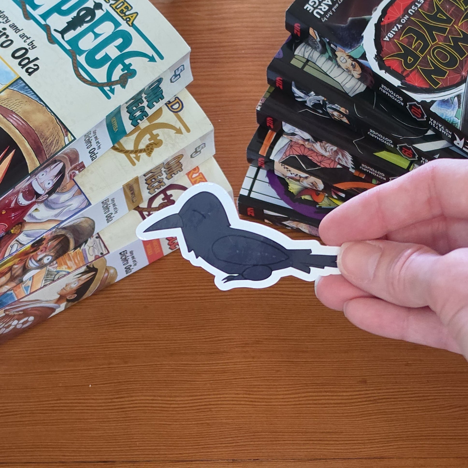 A hand holding a crow or raven sticker with a black body and white outline on a wooden table with stacks of manga books in the top-left and top-right.