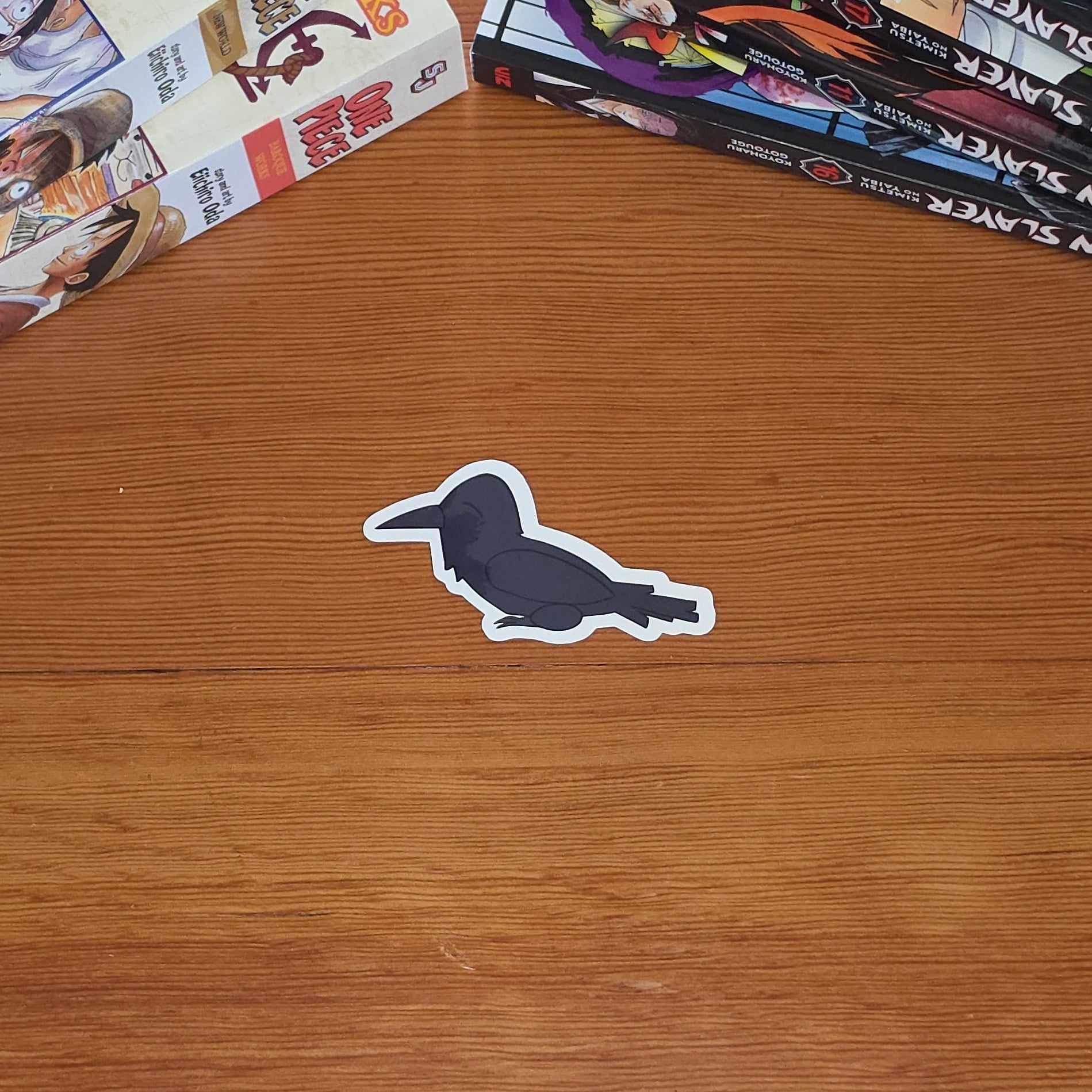 A close-up of a crow or raven sticker with a black body and white outline on a wooden table with stacks of manga books in the top-left and top-right.