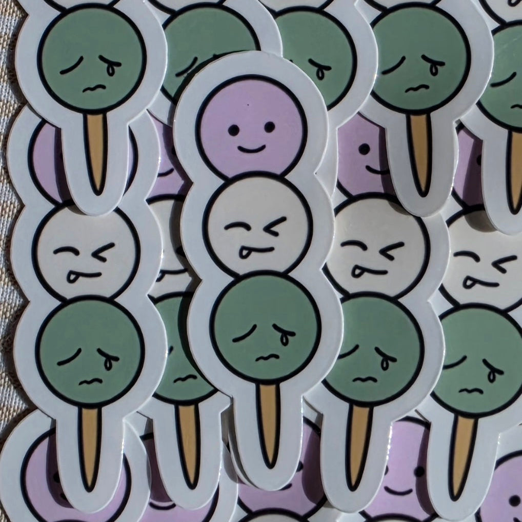 A close-up of multiple cartoon die-cut stickers arranged on a light surface. The stickers feature sad, crying faces with green, lavender, and white circular heads on tan sticks, outlined in black. Dango on a stick.