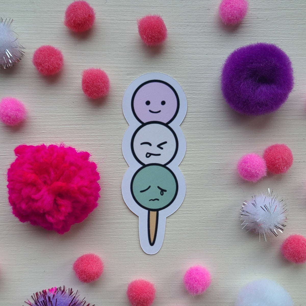 Sticker of three dango on a stick with colorful pom-poms on a light wooden surface.