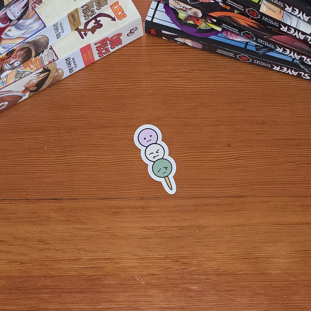 A small die-cut sticker featuring three stacked dango on a stick in white, light purple, and mint green, placed on a wooden tabletop next to stacks of manga books.